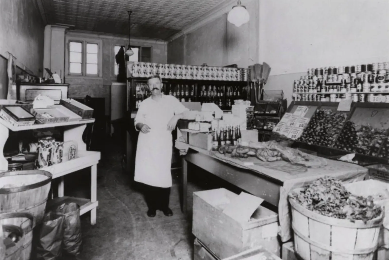 A grocer in a white apron standing in his shop among shelves of tins and baskets of produce